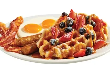 Denny’s Berry Waffle Slam featuring golden waffles topped with fresh strawberries, blueberries, whipped cream, and served with eggs and sausage.