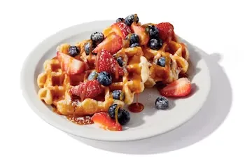Denny’s Berry Waffles topped with strawberries, blueberries, and whipped cream on a white plate.