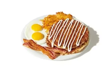 Denny’s Cinnamon Roll Pancake Slam with cinnamon swirl pancakes drizzled with icing, served with eggs, bacon, and hash browns.