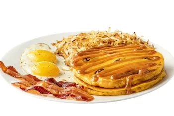Denny’s New Pumpkin Pecan Pancake Slam with fluffy pecan pancakes, crispy bacon, hash browns, and sunny-side eggs on a white plate.