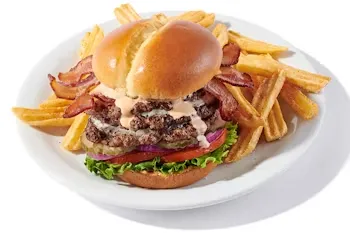 Denny’s Triple Juicy Burger stacked with beef patty, melted cheese, bacon, lettuce, tomato, and golden fries on a white plate.